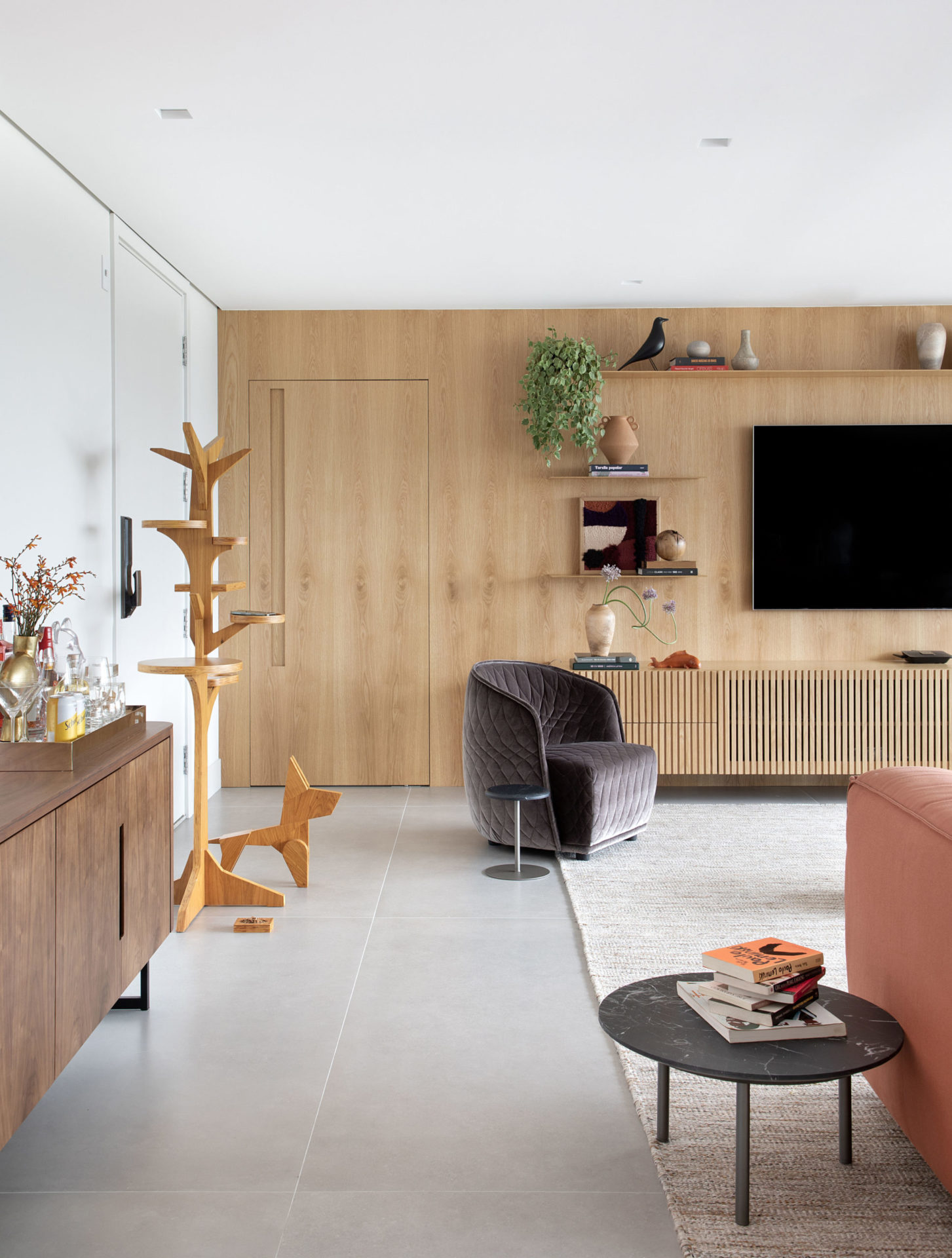 na foto há uma sala com TV na prede, um bufe, uma poltrona, uma mesinha de canto