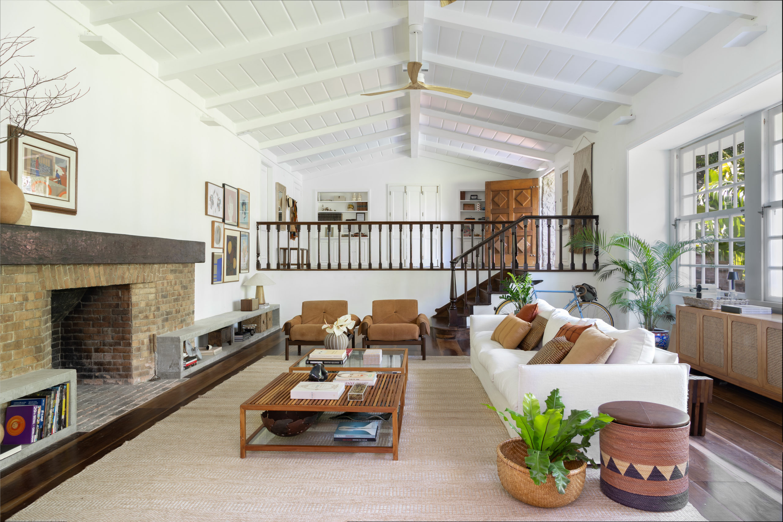 na foto imagem da sala com lareira, escada, sofá branco, puff e grandes janelas