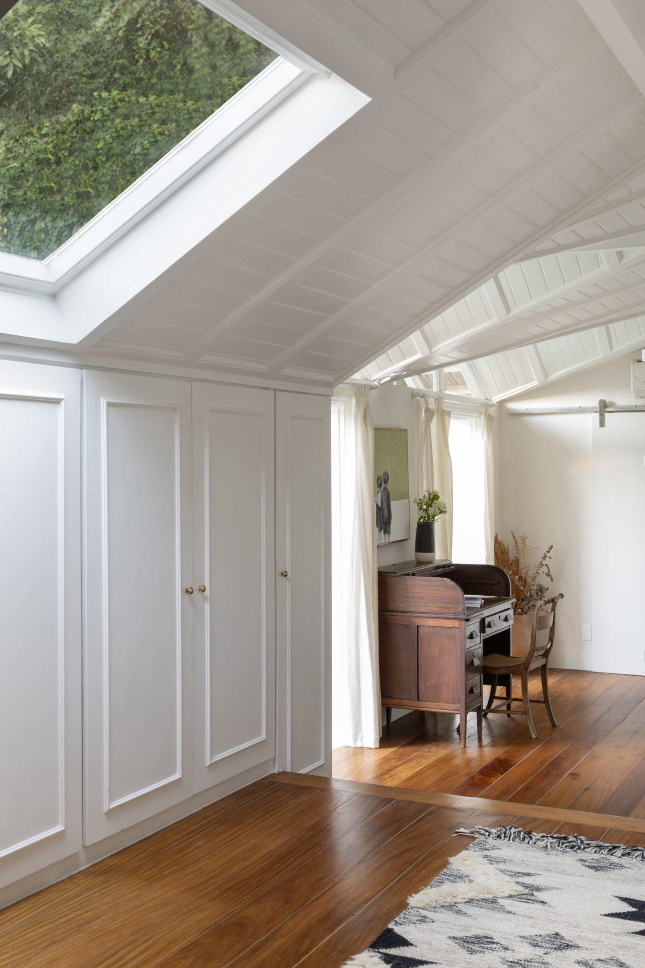 na foto quarto com claraboia, paredes brancas e uma escrivaninha
