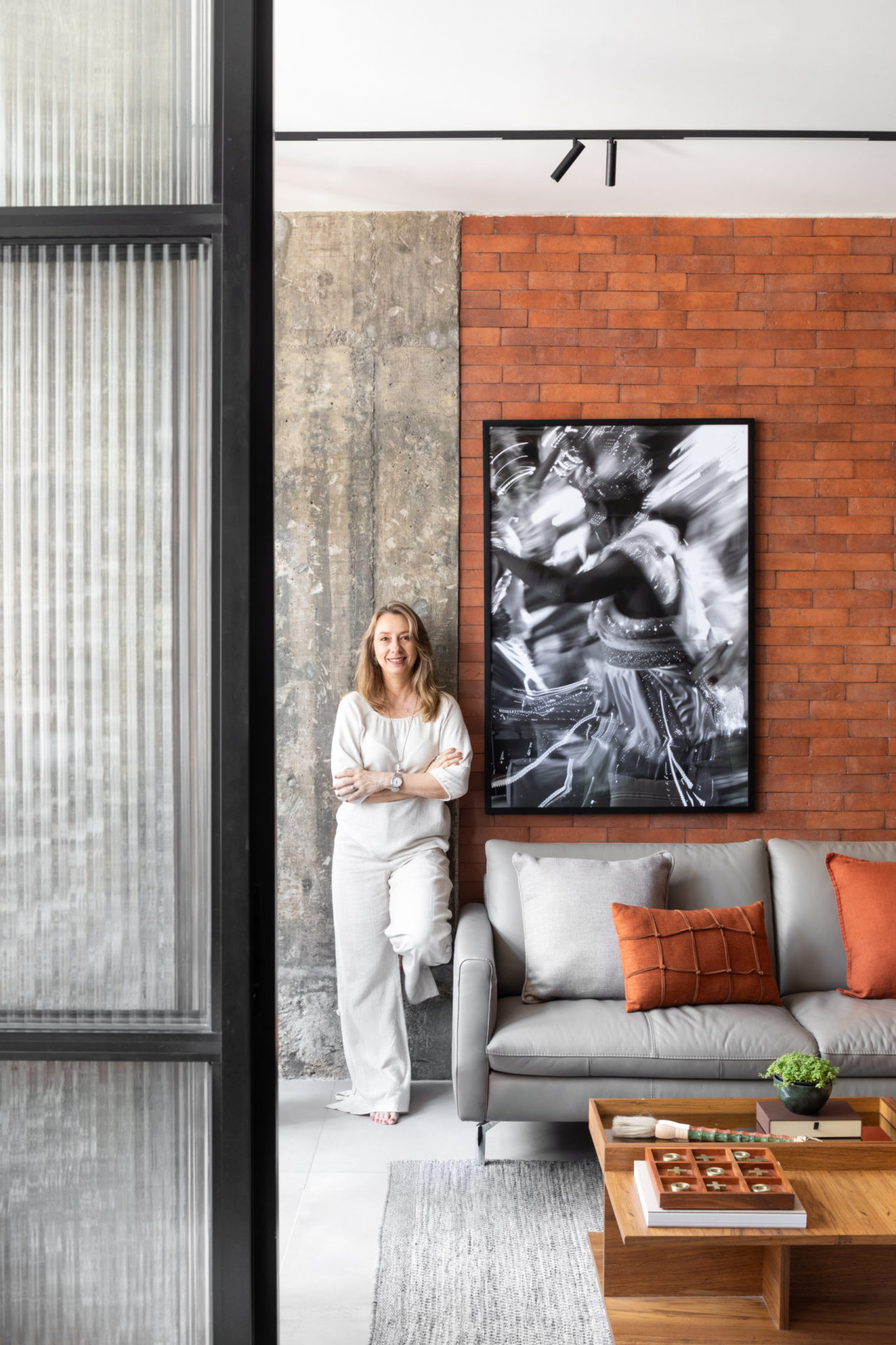 na foto uma mulher loira de cabelos longos em pé encostada na parede de tijolos de um loft