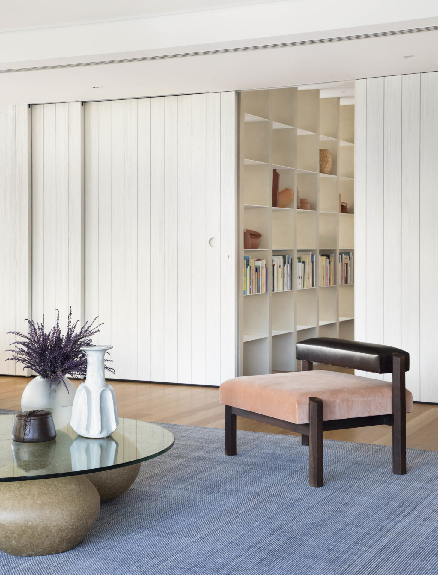 na foto parte do living com cadeira, mesa de centro de vidro e painel de lambri em madeira maciça ranhurada, pintada de branco