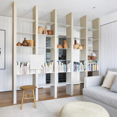 na foto sala com moveis claros, estante vasada e paredes brancas