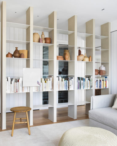 na foto sala com moveis claros, estante vasada e paredes brancas