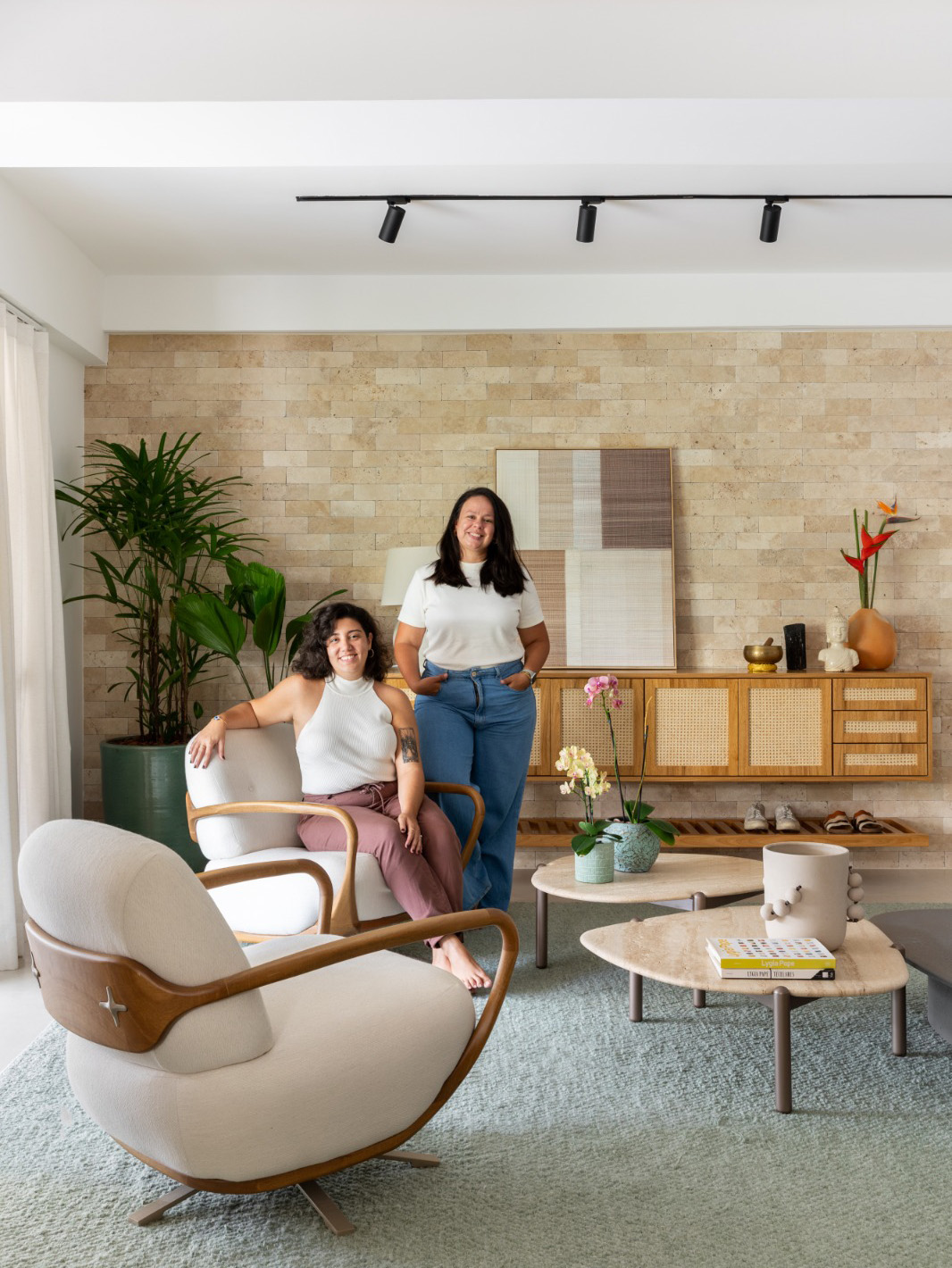 Duas mulheres brancas, uma sentada e outra em pé em uma sala de estar