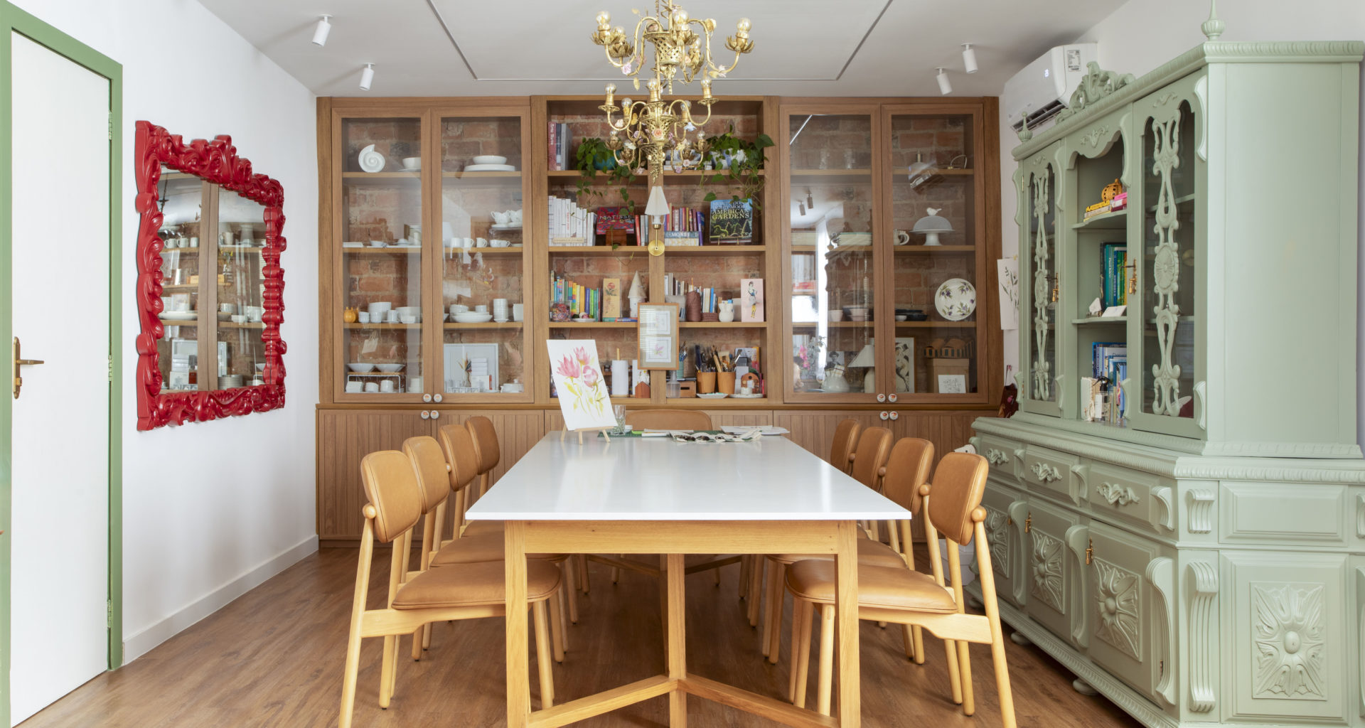 na foto mesa com seis cadeiras, duas estantes de madeira, lustre de ferro e espelho com moldura vermelha