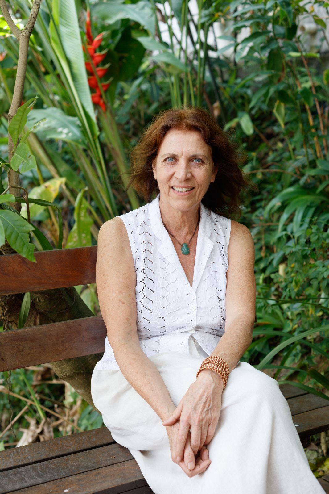 na foto mulher branca, de cabelos ruivos, vestindo roupa branca sentada em um banco de madeira. 