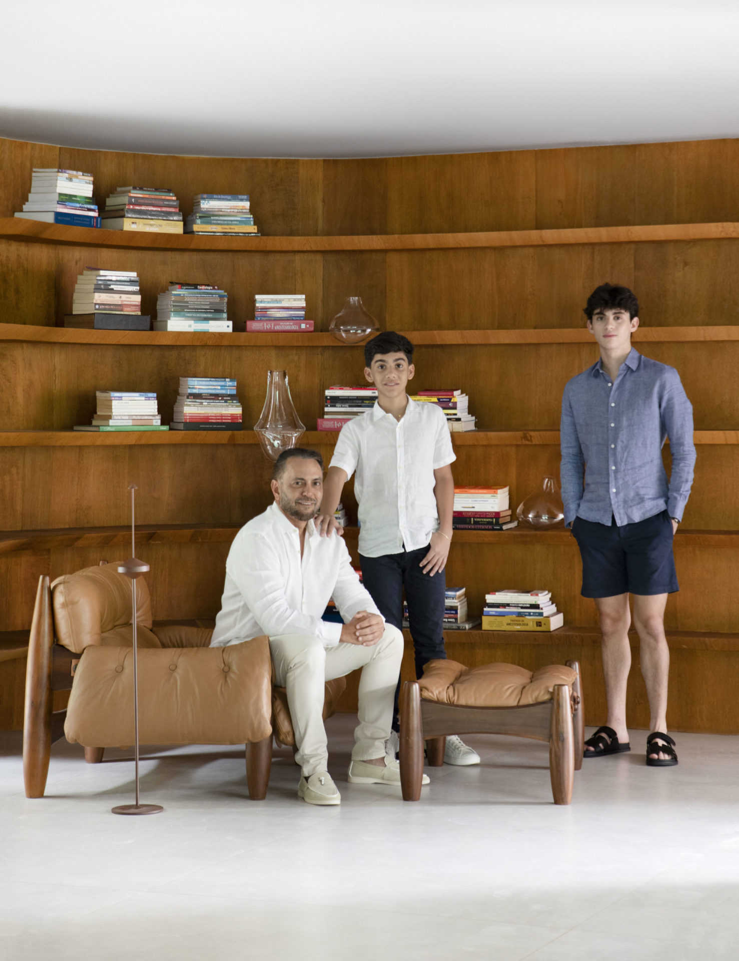 na foto um homem sentado e dois meninos em pé. Atrás uma estante de madeira com livros