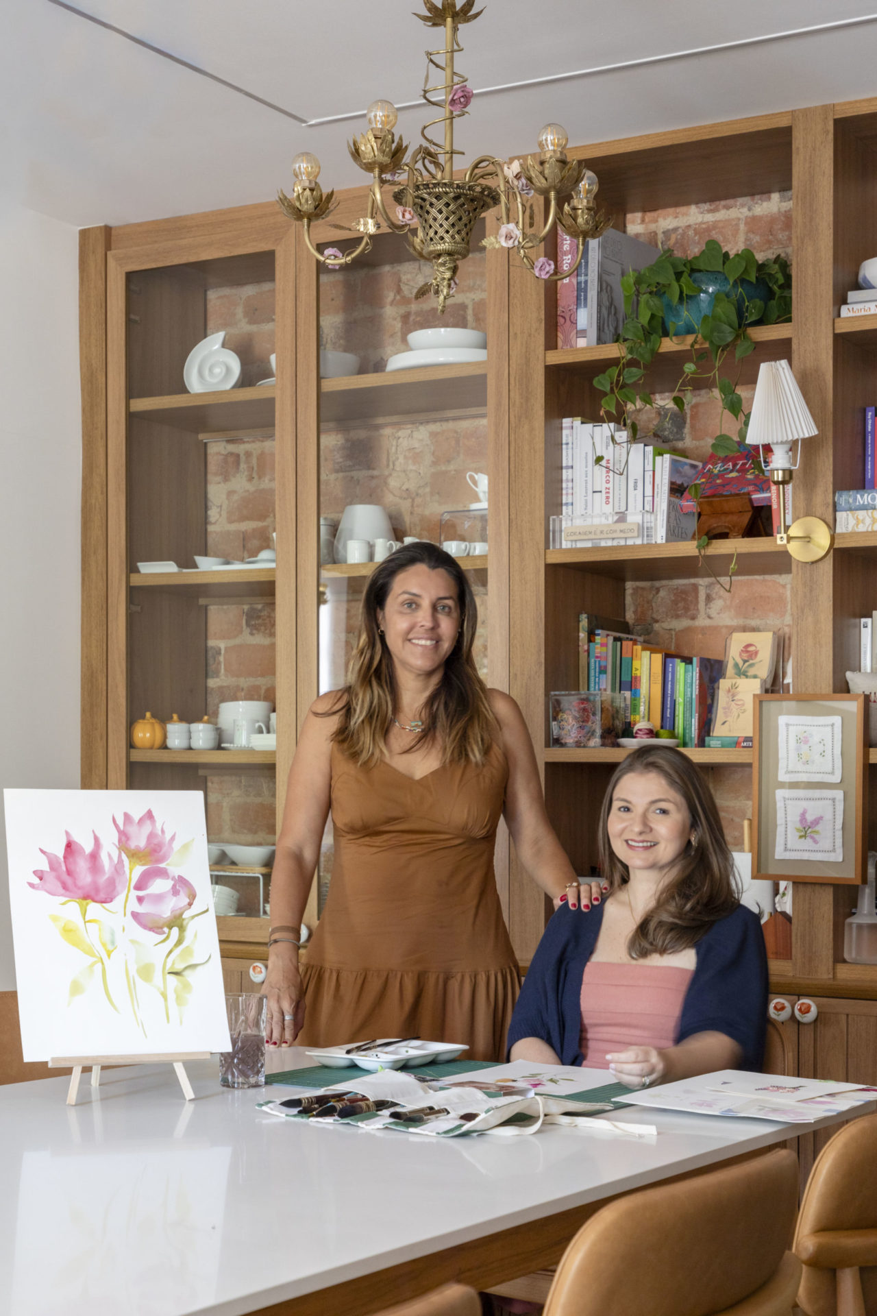 na foto uma mulher em pé e uma mulher sentada à mesa de um atelier