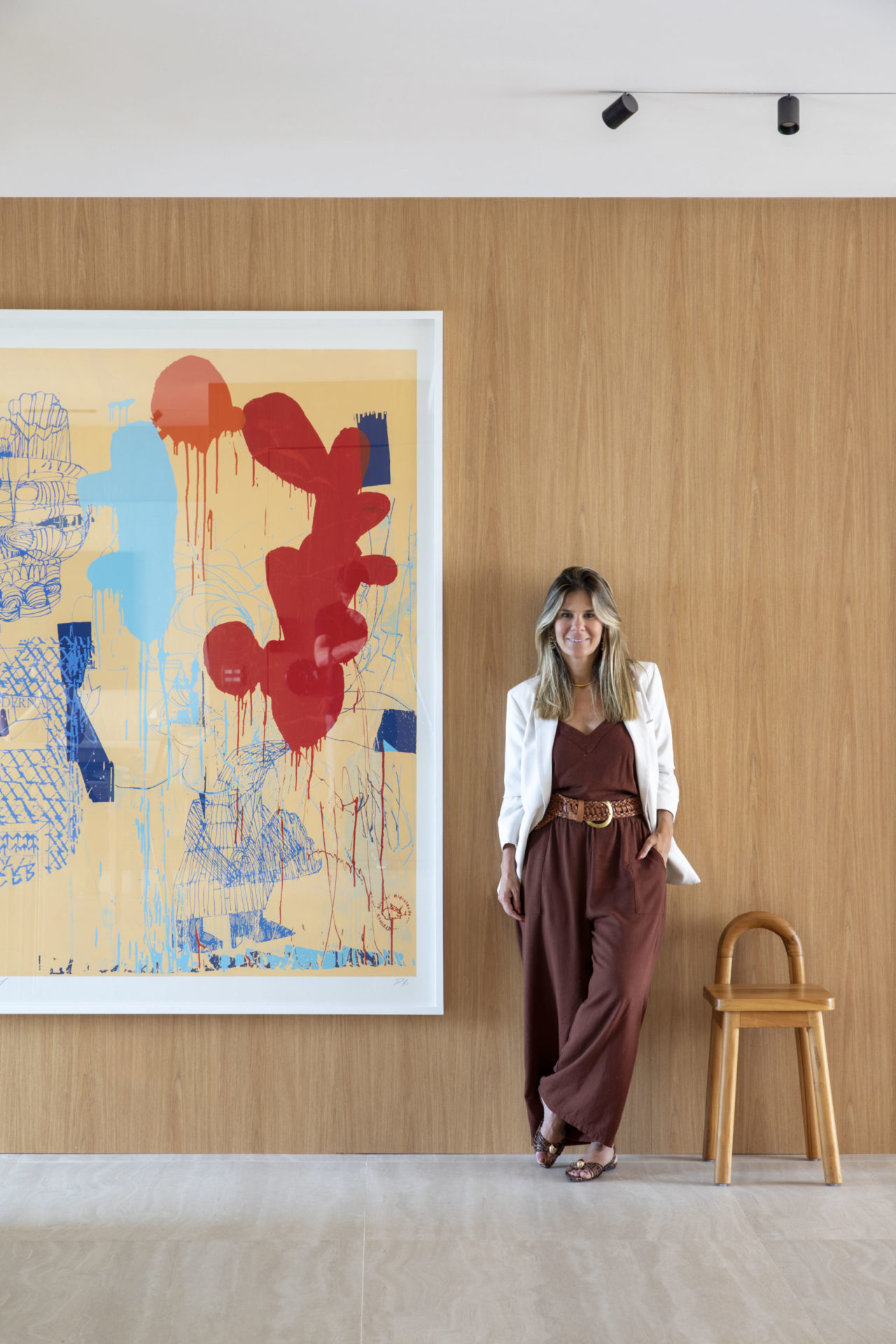 na foto mulher loura, de roupa marrom e branca encostada na parede onde há um quadro grande e colorido