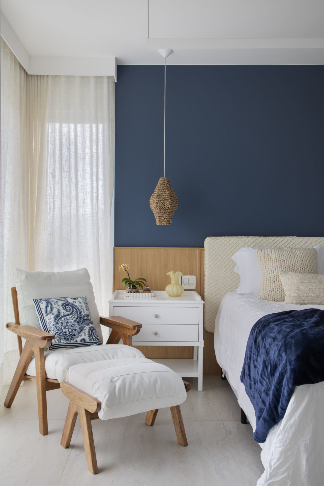 na foto quarto com cama branca, espreguiçadeira de madeira e tecido branco e parede azul