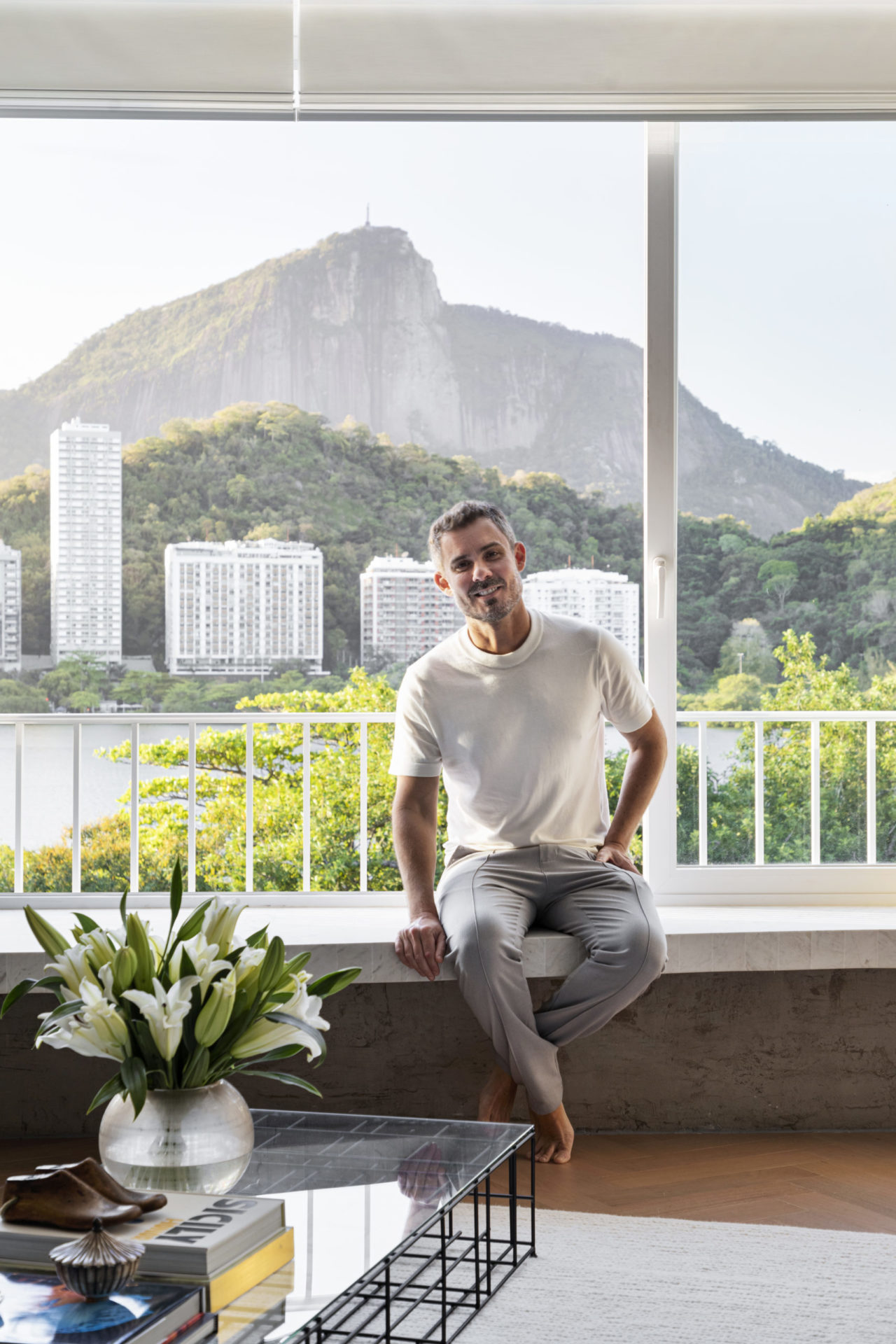 na foto home branco com cala jeans, blusa branca sentado em janela com vista para a Lagoa Rodrigo de Freitas