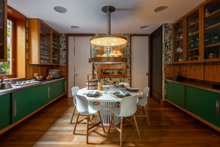 Copa de uma cozinha bem clara. Mesa redonda branca, central. Armarios superiores em madeira, e os da parte de baixo, na cor verde. 