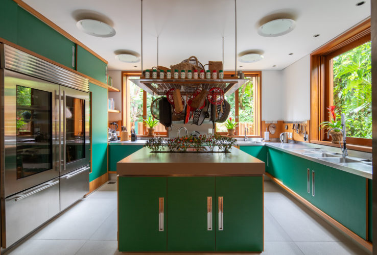 Cozinha clara, com luz natural. Bancada em inox, janelas em madeira e os armarios, na cor verde. Uma ilha central com pendurador de panelas. 