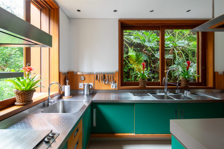 Cozinha clara, com luz natural. Bancada em inox, janelas em madeira e os armarios, na cor verde.