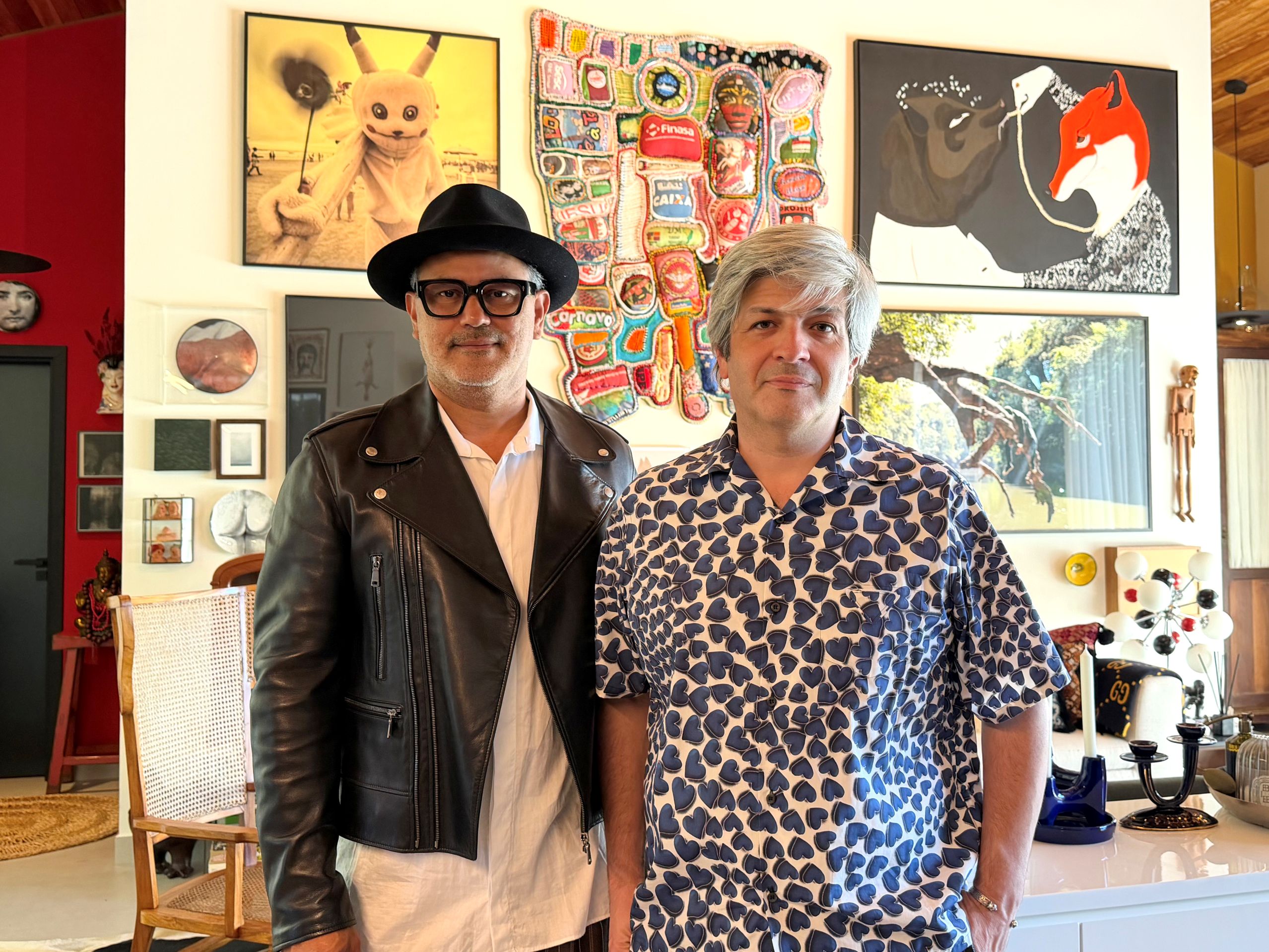 Na foto dois homens brancos. Um com chapéu preto e muitas obras de arte atrás deles