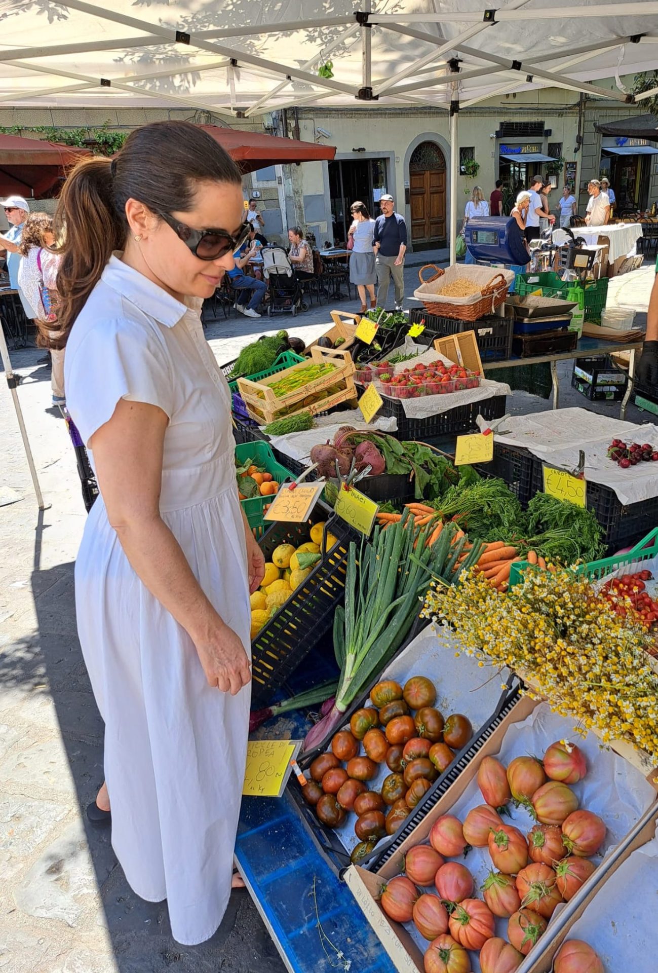 na foto uma feira com alimentos e mulher branca de óculos escuros olhando os produtos