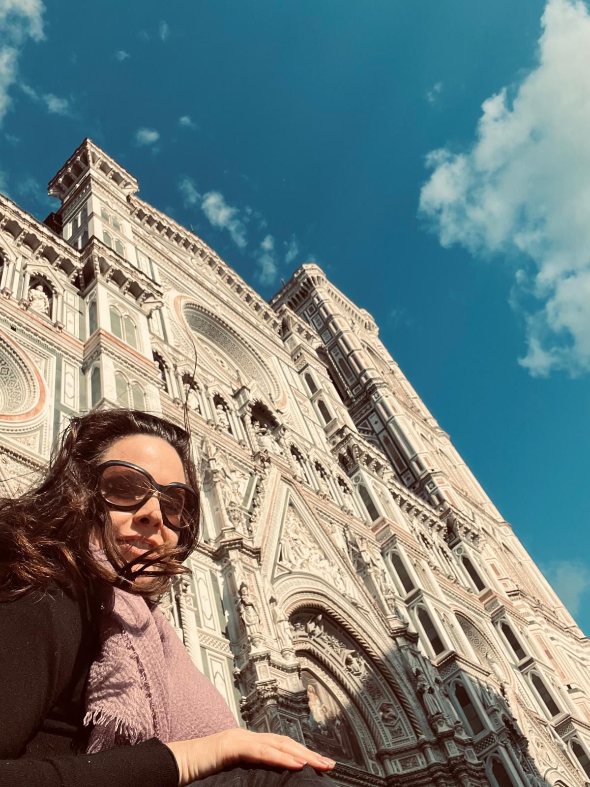 na foto mulher branca e com óculos escuros e um castelo atrás. céu azul com nuvens