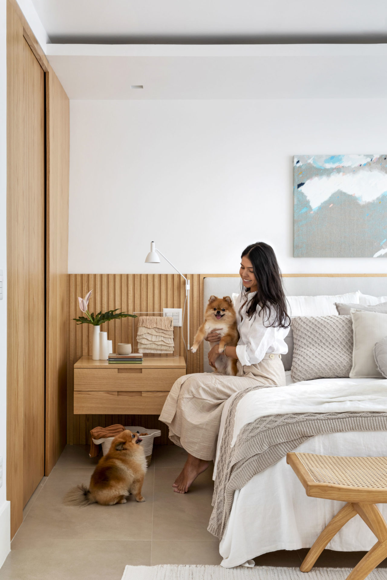 na foto um quarto de casal com uma mulher sentada na cama com dois cachorrinhos