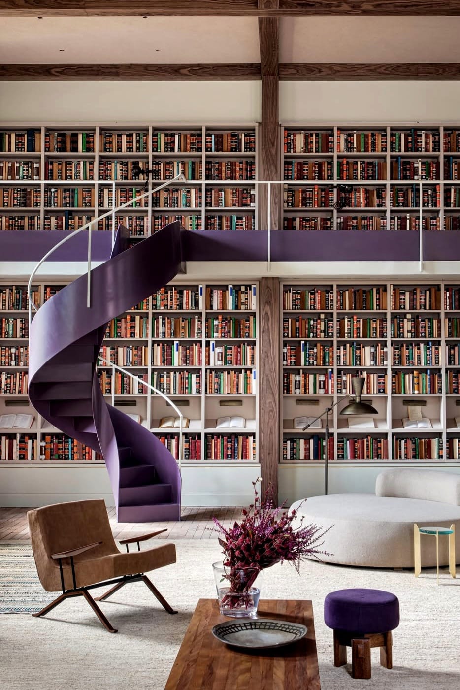na foto espaço com grande estante com livros, uma escada em tom lavanda, uma cadeira e mesa