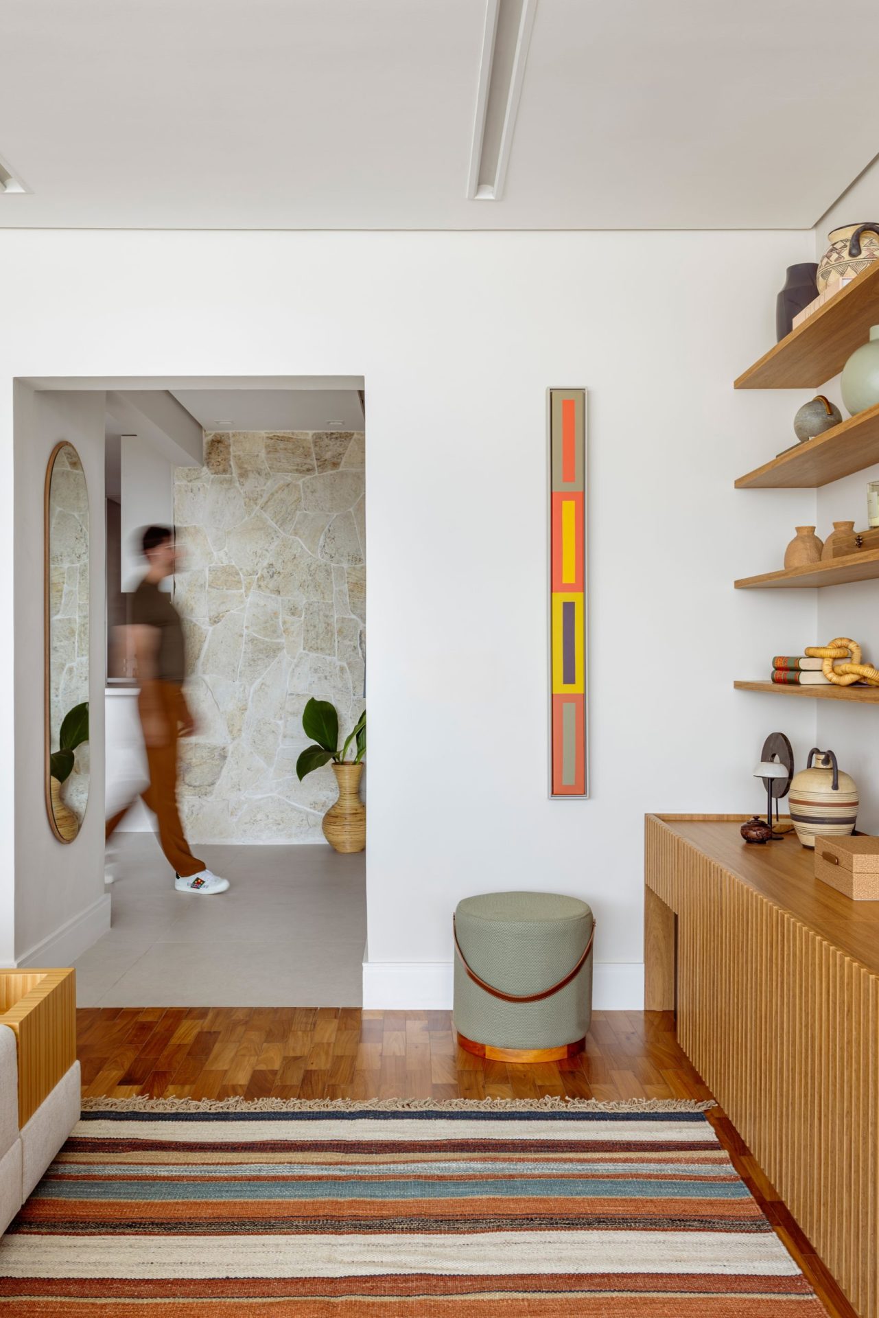 na foto interior de apartamento com tapete colorido, estante de madeira. na foto um homem passa pelo corredor